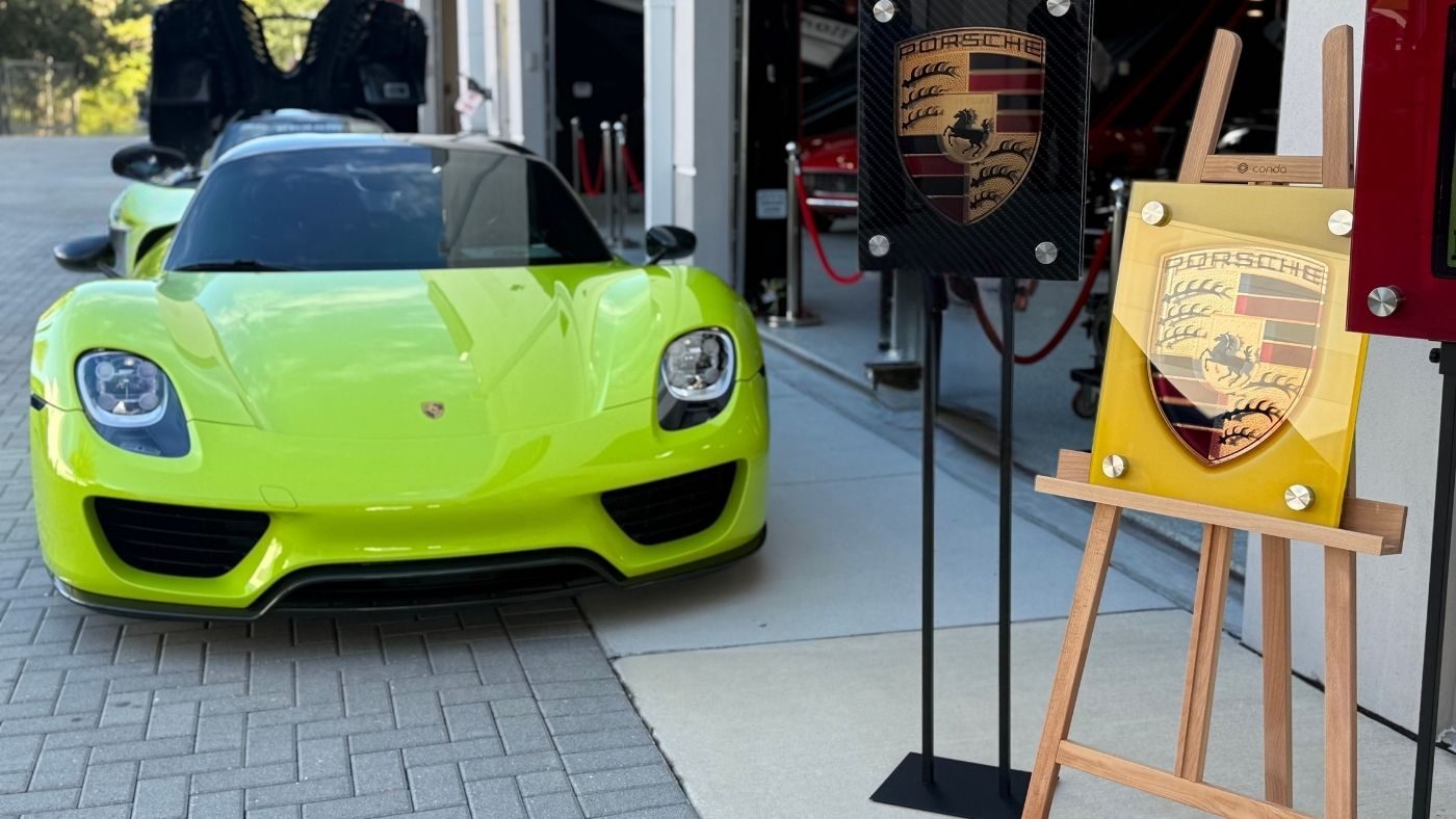 Illuminated glass Porsche crest art on a stand beside a Porsche 918 by Dyer Design Glassworks
